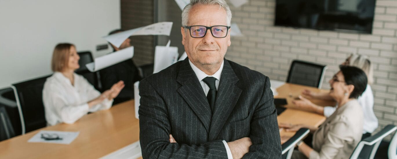 Business executive standing confidently in meeting room with team engaged in discussion behind.