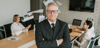 Business executive standing confidently in meeting room with team engaged in discussion behind.