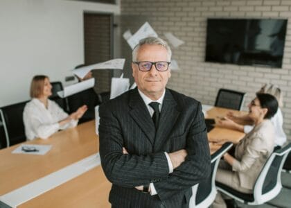 Business executive standing confidently in meeting room with team engaged in discussion behind.